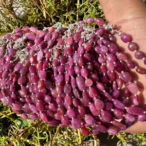 May include: A close-up of a pile of pink ruby gemstone bracelets with silver clasps. The bracelets are made of oval-shaped beads and are strung on silver chains.