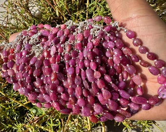 May include: A close-up of a pile of pink ruby gemstone bracelets with silver clasps. The bracelets are made of oval-shaped beads and are strung on silver chains.