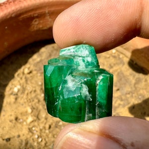 May include: A large, green emerald crystal with a rough, natural texture. The crystal has a deep green color and is partially covered in white inclusions.