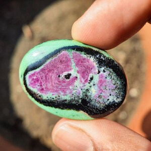 May include: Oval-shaped gemstone with a vibrant color palette. The stone features a central area of swirling magenta, surrounded by a light green band, a black border, and a green outer edge. The polished surface reflects light.