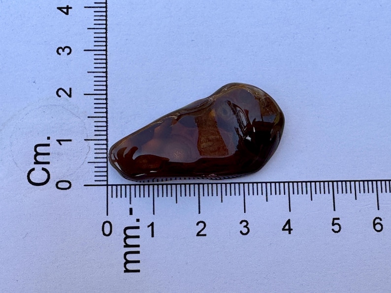 May include: A polished, dark brown, irregular-shaped stone cabochon with a smooth, glossy surface. The stone is positioned against a ruler, showing a length of approximately 3.5 centimeters and a width of approximately 1.5 centimeters.