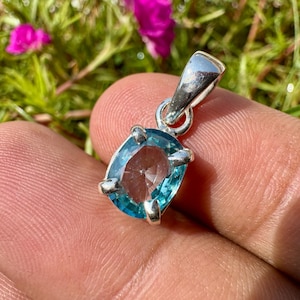 May include: A silver pendant featuring a faceted, oval-shaped blue gemstone held by four prongs. The pendant includes a polished silver bail at the top for attachment, showcasing a clear, vibrant blue color and a reflective, multi-faceted surface.