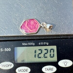 K&ouml;nnte beinhalten: Ein silberner Anh&auml;nger mit einem rosa Stein mit hexagonalem Muster, der auf einer digitalen Waage liegt, die ein Gewicht von 12,20 ct anzeigt. Der Anh&auml;nger hat eine polierte silberne &Ouml;se und einen glatten, unregelm&auml;&szlig;igen Metallrahmen.