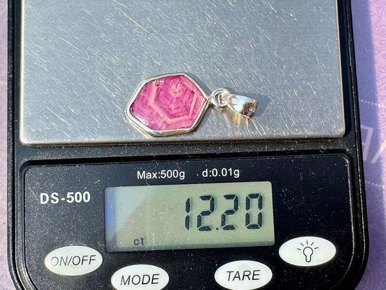 K&ouml;nnte beinhalten: Ein silberner Anh&auml;nger mit einem rosa Stein mit hexagonalem Muster, der auf einer digitalen Waage liegt, die ein Gewicht von 12,20 ct anzeigt. Der Anh&auml;nger hat eine polierte silberne &Ouml;se und einen glatten, unregelm&auml;&szlig;igen Metallrahmen.