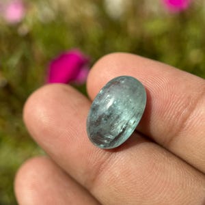 May include: A smooth, oval-shaped aquamarine gemstone with a light blue-green colour. The stone has a slightly cloudy appearance and is set in a cabochon cut.