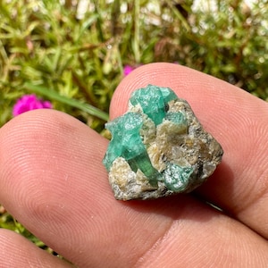 以下が含まれることがあります： 明るい緑色の六角柱状の結晶が、質感のある薄茶色と灰色の岩石母岩に埋め込まれた、原石のエメラルド標本。この鉱物クラスターは、半透明の緑色の面と、表面に自然な結晶構造を示しています。