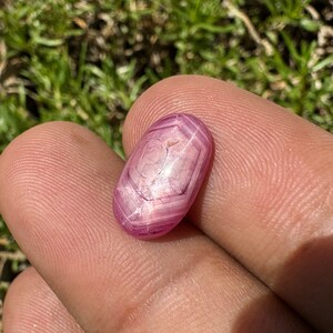 May include: A pink star ruby gemstone with a hexagonal pattern. The gemstone is cut into an oval shape and has a polished finish.