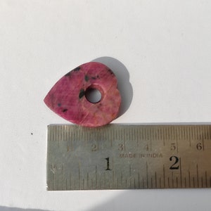 May include: A pink heart-shaped pendant with a hole in the center. The pendant is approximately 1 inch in length and is made of a pink and black stone. The pendant is sitting on a ruler with the measurement of 1 inch.