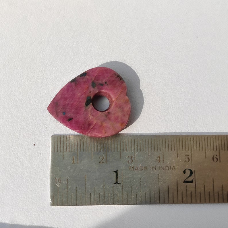 May include: A pink heart-shaped pendant with a hole in the center. The pendant is approximately 1 inch in length and is made of a pink and black stone. The pendant is sitting on a ruler with the measurement of 1 inch.