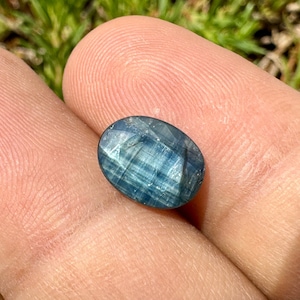 May include: An oval-shaped, faceted gemstone with a blue hue. The gem has a polished surface with visible internal striations. The stone is held in a hand, with a blurred green background.