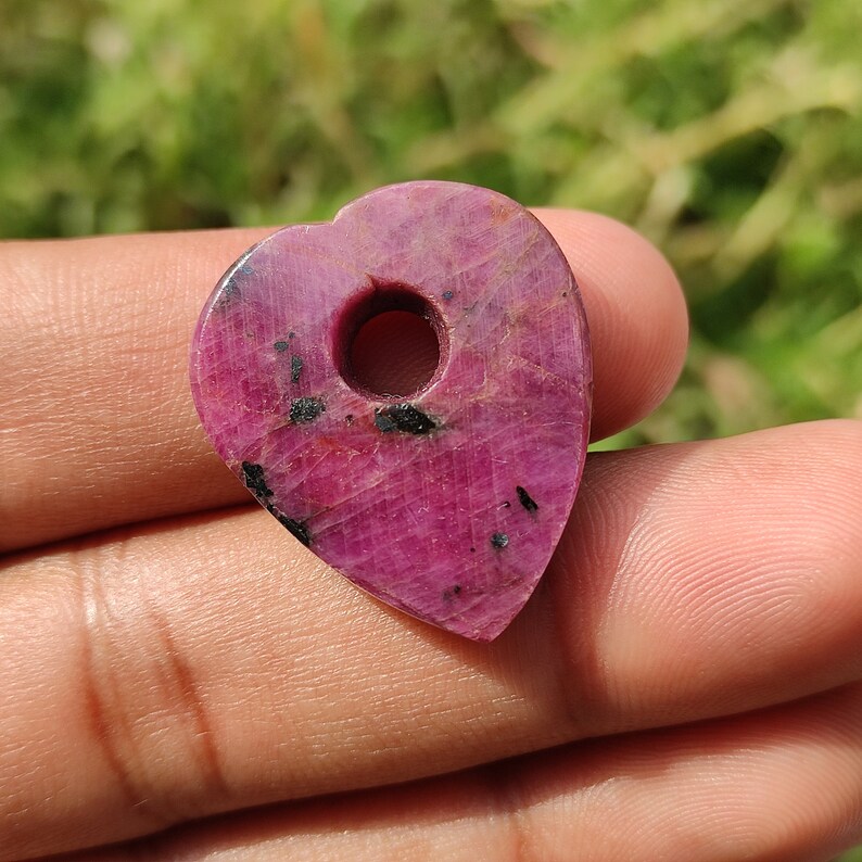 May include: A pink heart-shaped pendant made of ruby with black inclusions. The pendant has a hole drilled through the top for stringing.