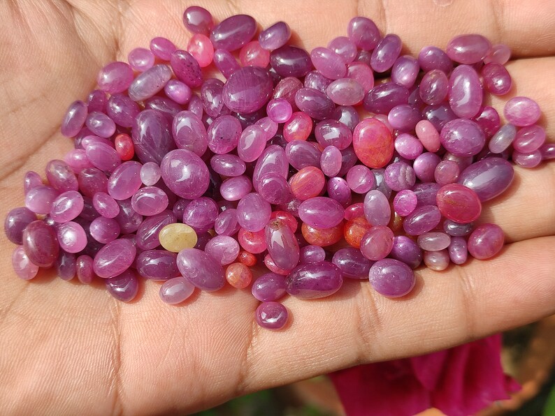 May include: A handful of polished, oval-shaped gemstones in shades of pink and red. The gemstones vary in size and have a smooth, glossy surface. A few stones have a yellowish hue.