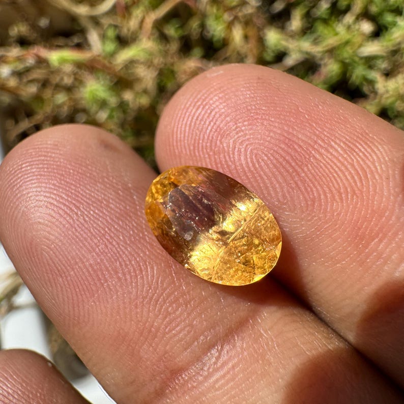 May include: A golden yellow oval-shaped gemstone with a slight reddish hue and visible internal inclusions.