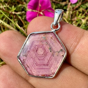 May include: A hexagonal pink tourmaline pendant set in a polished silver frame. The gemstone features distinct concentric hexagonal growth patterns in shades of light and dark pink, with small natural inclusions visible near the top edge.