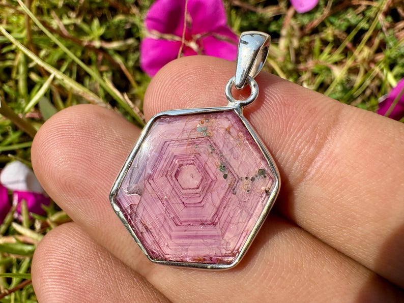 K&ouml;nnte beinhalten: Ein sechseckiger rosa Turmalin-Anh&auml;nger in einer polierten Silberfassung. Der Edelstein weist ausgepr&auml;gte konzentrische hexagonale Wachstumsmuster in hellen und dunklen Rosat&ouml;nen auf, mit kleinen nat&uuml;rlichen Einschl&uuml;ssen am oberen Rand.