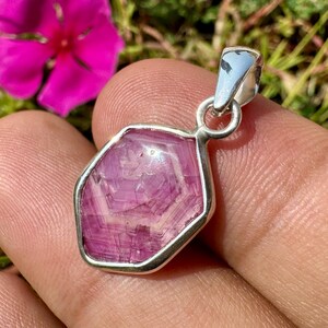 K&ouml;nnte beinhalten: Ein Anh&auml;nger mit einem facettierten, sechseckigen rosa Edelstein in einer Fassung aus poliertem silberfarbenem Metall. Der Stein zeigt interne kristalline Muster und Lichtreflexionen und ist an einer silberfarbenen Metall&ouml;se befestigt.