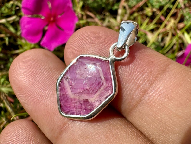 K&ouml;nnte beinhalten: Ein Anh&auml;nger mit einem facettierten, sechseckigen rosa Edelstein in einer Fassung aus poliertem silberfarbenem Metall. Der Stein zeigt interne kristalline Muster und Lichtreflexionen und ist an einer silberfarbenen Metall&ouml;se befestigt.