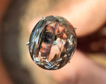 May include: Oval-shaped gemstone with a faceted surface, showcasing a blend of clear, brown, and iridescent colors. The gem is held by a silver setting. The stone's internal structure displays linear patterns, creating a unique visual effect.