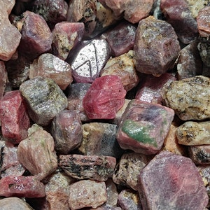 Puede incluir: Primer plano de un montón de piedras preciosas de rubí en bruto, de color rosa y rojo. Las piedras son de varios tamaños y formas, algunas muestran un tono verde.