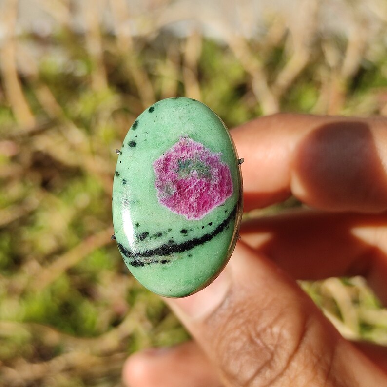 Peut inclure: Pierre pr&eacute;cieuse verte de forme ovale avec des inclusions noires et blanches et un centre rose. La pierre pr&eacute;cieuse est sertie dans une b&eacute;li&egrave;re en argent.
