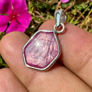 May include: A hexagonal pink gemstone pendant set in a polished silver frame. The stone features visible internal striations and a translucent quality, held by a silver bail at the top, showcasing a geometric shape and natural crystalline texture.