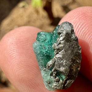 以下が含まれることがあります： 鮮やかで半透明な緑色のエメラルドの結晶と、銀色の金属光沢を持つパイライトの塊が融合した原石の標本。滑らかな緑色の結晶と、ギザギザとした反射性のあるパイライトの金属表面との間で、質感の対比が見られる自然で不規則な形状の石です。