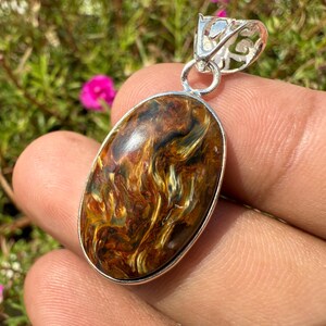 May include: Oval pendant with a swirling pattern of brown, gold, and black hues. The gemstone is set in a silver-colored metal frame with a decorative bail. The pendant is held in a hand against a backdrop of green foliage and pink flowers.