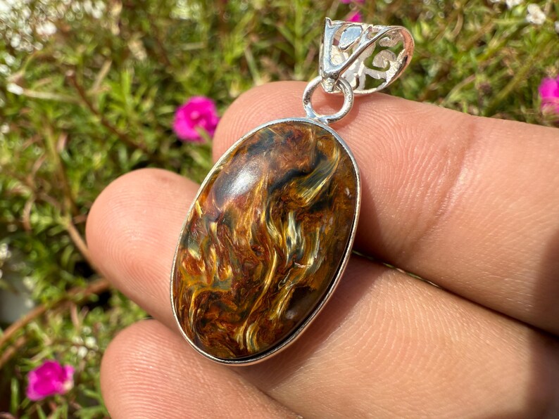 May include: Oval pendant with a swirling pattern of brown, gold, and black hues. The gemstone is set in a silver-colored metal frame with a decorative bail. The pendant is held in a hand against a backdrop of green foliage and pink flowers.