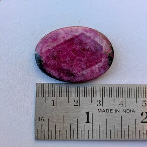 May include: Oval-shaped, faceted ruby gemstone with a deep magenta hue. The gemstone is set against a white surface, with a metal ruler showing measurements. The ruby has a polished surface and a dark border.