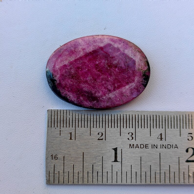 May include: Oval-shaped, faceted ruby gemstone with a deep magenta hue. The gemstone is set against a white surface, with a metal ruler showing measurements. The ruby has a polished surface and a dark border.