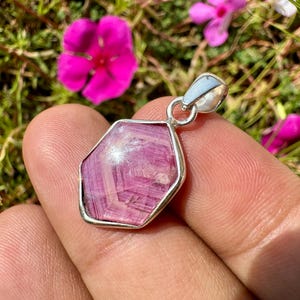 K&ouml;nnte beinhalten: Ein sechseckiger Anh&auml;nger mit einem facettierten rosa Edelstein, der in eine polierte silberfarbene Metallfassung eingefasst ist und &uuml;ber eine passende silberfarbene &Ouml;se zur Befestigung verf&uuml;gt.