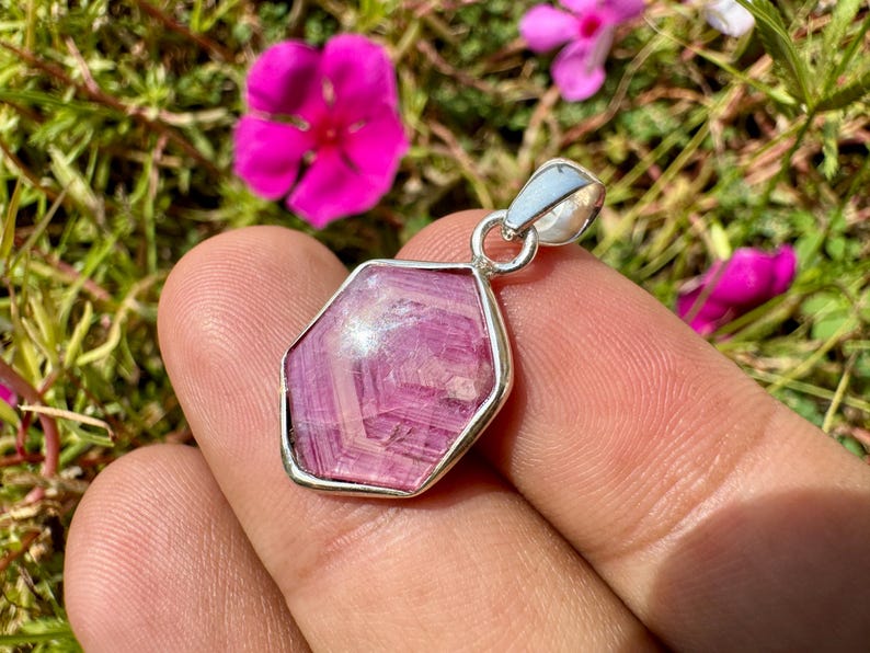 K&ouml;nnte beinhalten: Ein sechseckiger Anh&auml;nger mit einem facettierten rosa Edelstein, der in eine polierte silberfarbene Metallfassung eingefasst ist und &uuml;ber eine passende silberfarbene &Ouml;se zur Befestigung verf&uuml;gt.