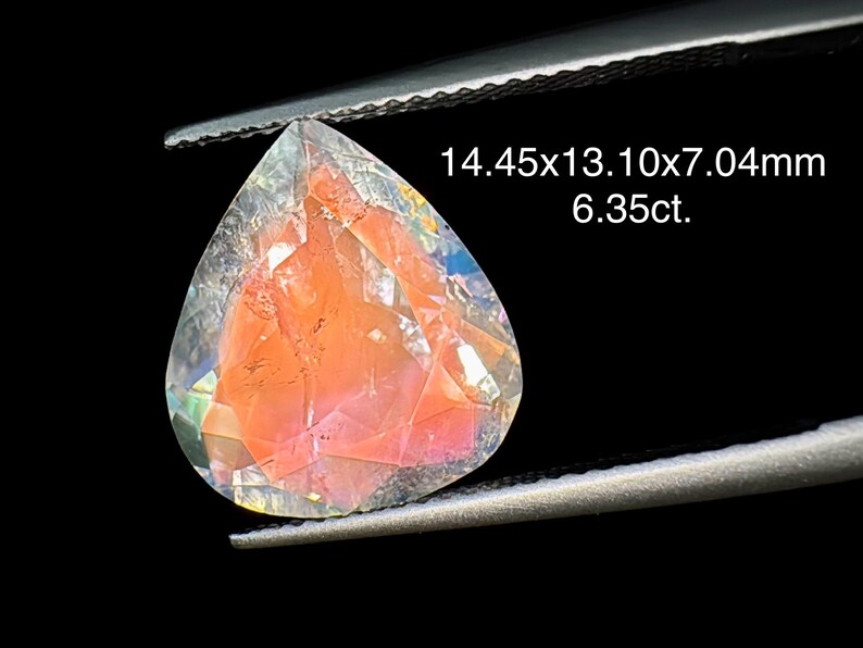 May include: A clear, faceted gemstone with a pink hue and iridescent rainbow colors. The gemstone is shaped like a teardrop and measures 14.45x13.10x7.04mm, weighing 6.35 carats.