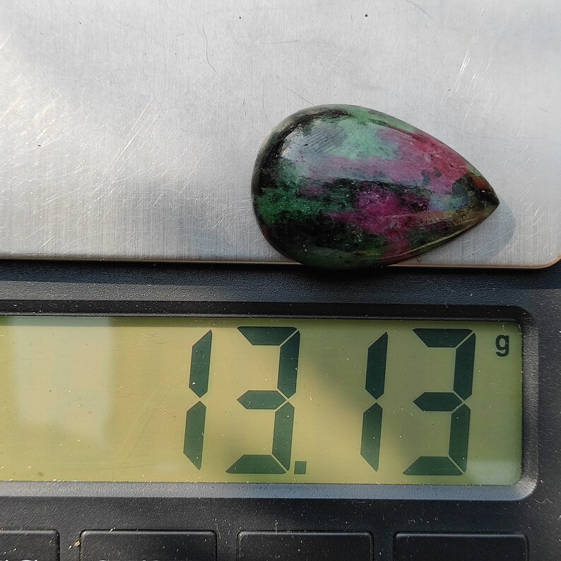 May include: A green, pink, and black gemstone cabochon weighing 13.13 grams on a digital scale.