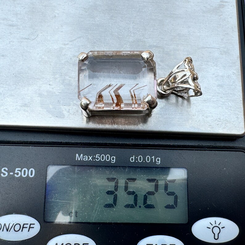 K&ouml;nnte beinhalten: Ein klarer Quarz-Anh&auml;nger mit braunen Einschl&uuml;ssen, in Silber gefasst, wiegt 3,25 Gramm. Der Anh&auml;nger befindet sich auf einer digitalen Waage.