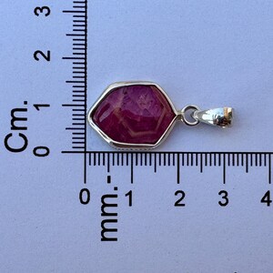 K&ouml;nnte beinhalten: Ein sechseckiger Anh&auml;nger mit einem tief magentafarbenen Edelstein in einer silberfarbenen Metallfassung. Der Anh&auml;nger liegt neben einem Lineal, das eine Breite von etwa 1,9 cm und eine H&ouml;he von 1,3 cm anzeigt.