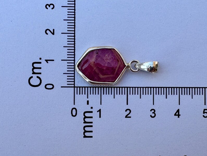 K&ouml;nnte beinhalten: Ein sechseckiger Anh&auml;nger mit einem tief magentafarbenen Edelstein in einer silberfarbenen Metallfassung. Der Anh&auml;nger liegt neben einem Lineal, das eine Breite von etwa 1,9 cm und eine H&ouml;he von 1,3 cm anzeigt.