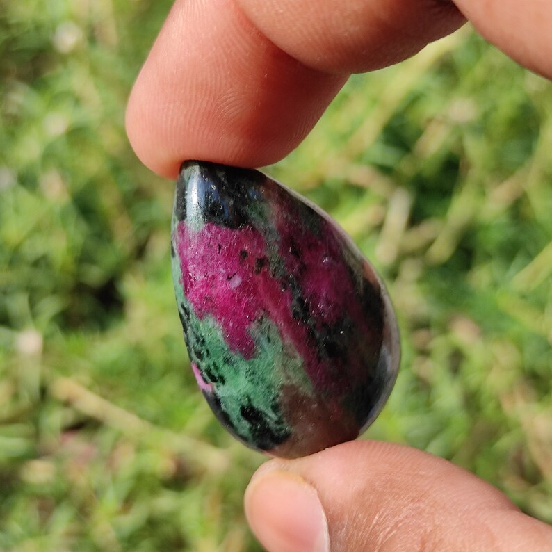 May include: A teardrop-shaped green, pink, and black gemstone pendant. The gemstone has a mottled appearance with swirls of color.