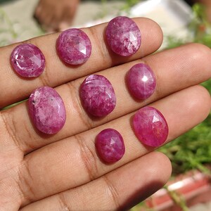 Pu&ograve; includere: Una collezione di nove pietre preziose di rubino sfaccettate di forma ovale in diverse tonalit&agrave; di rosa. Le pietre preziose sono esposte su una mano.