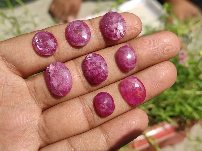 Pu&ograve; includere: Una collezione di nove pietre preziose di rubino sfaccettate di forma ovale in diverse tonalit&agrave; di rosa. Le pietre preziose sono esposte su una mano.