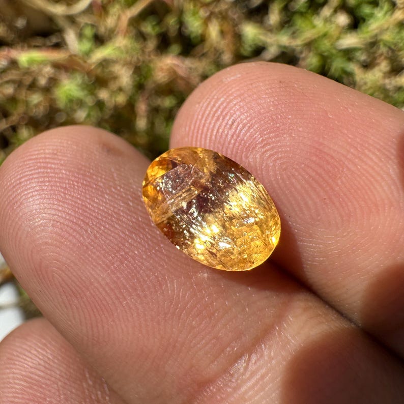 May include: A golden yellow oval-shaped gemstone with a smooth, polished surface. The stone has a translucent appearance with visible internal striations.