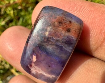 May include: A rectangular gemstone with a polished surface. The stone features a blend of colors, including deep purple, reddish-brown, and hints of blue and white. The gemstone is held in a hand, with a blurred green background.
