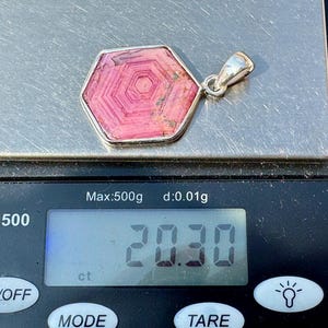 K&ouml;nnte beinhalten: Ein sechseckiger, rosafarbener Rhodochrosit-Anh&auml;nger mit einer silberfarbenen &Ouml;se, der auf einer digitalen Waage mit einem Gewicht von 20,30 ct liegt. Der Stein zeigt konzentrische, sechseckige Streifenmuster in Rosa- und Pinkt&ouml;nen.