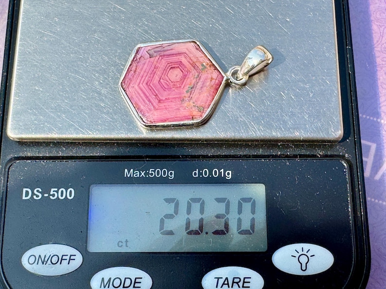 K&ouml;nnte beinhalten: Ein sechseckiger, rosafarbener Rhodochrosit-Anh&auml;nger mit einer silberfarbenen &Ouml;se, der auf einer digitalen Waage mit einem Gewicht von 20,30 ct liegt. Der Stein zeigt konzentrische, sechseckige Streifenmuster in Rosa- und Pinkt&ouml;nen.