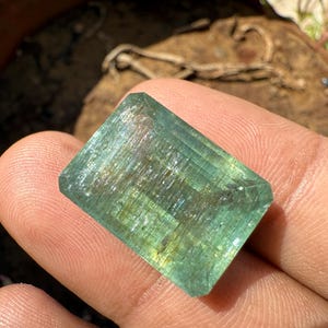 Op de afbeelding: Een rechthoekige, gefacetteerde edelsteen met een lichtgroene en blauwe kleur. De edelsteen wordt in een hand gehouden en de facetten vangen het licht op, waardoor een glinsterend effect ontstaat. De edelsteen is een natuurlijk mineraal, waarschijnlijk voor sieraden.