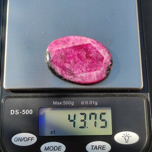 May include: A pink oval gemstone on a digital scale, displaying a weight of 43.75 ct. The gemstone has a lighter pink center and a darker pink border, with black accents. The scale reads 'Max:500g d:0.01g'.