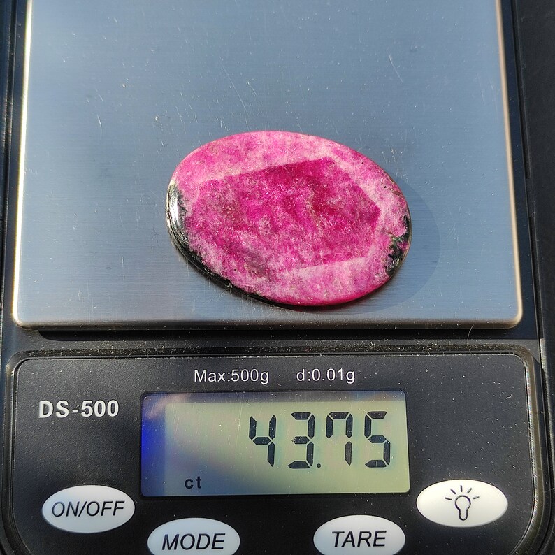 May include: A pink oval gemstone on a digital scale, displaying a weight of 43.75 ct. The gemstone has a lighter pink center and a darker pink border, with black accents. The scale reads 'Max:500g d:0.01g'.