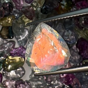 May include: A clear, faceted gemstone with a pink interior. The gemstone is held by tweezers and surrounded by other loose gemstones.