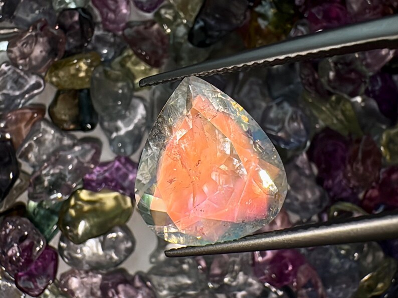 May include: A clear, faceted gemstone with a pink interior. The gemstone is held by tweezers and surrounded by other loose gemstones.