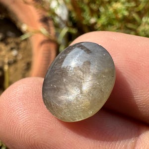 以下が含まれることがあります： 光沢のある虹色の表面を持つ、銀灰色の楕円形の宝石。石は滑らかで磨かれており、控えめな光沢があります。